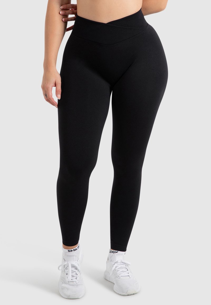 Zwarte high-waisted leggings met een gladde textuur en een crossover taillebandontwerp, gecombineerd met witte sportschoenen.