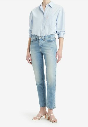 Personne portant une chemise bleu clair boutonnée, un jean bleu slim délavé et des sandales beiges à brides avec petits talons blocs, debout sur un fond blanc.