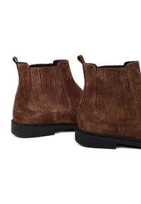 Stivaletti in suede marrone con una texture liscia, pannelli laterali elastici e una suola in gomma nera. Cuciture dettagliate sul retro.