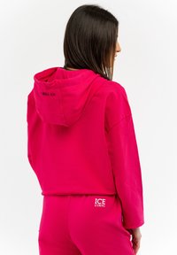 Sudadera rosa con cordón y logo bordado "ICE" en la parte posterior. La tela parece suave, con mangas largas y un corte relajado.
