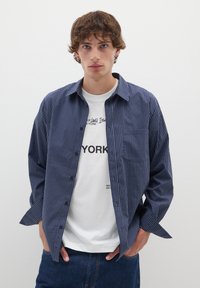 Camicia a quadri blu navy con bottoni e una tasca, indossata sopra una T-shirt bianca con stampa. Pantaloni in denim. Sfondo semplice.