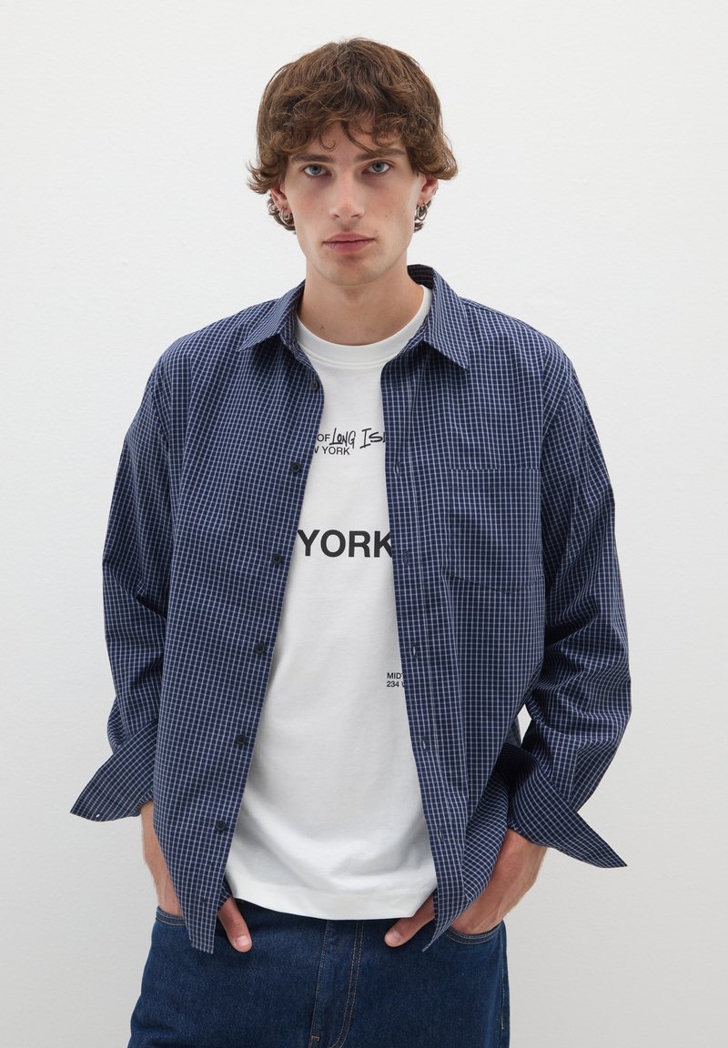 Camicia a quadri blu navy con bottoni e una tasca, indossata sopra una T-shirt bianca con stampa. Pantaloni in denim. Sfondo semplice.