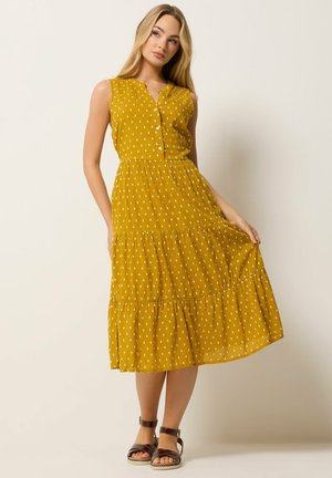 Frau in ärmellosem midilangen senfgelben Kleid mit kleinen weißen Punkten, mit Knopfleiste vorne, hält den Saum, trägt braune Sandalen, steht vor neutralem Hintergrund.