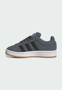 Zapatilla de gamuza gris con tres franjas negras, suela de goma blanca y suela de goma natural. Punta redonda y detalle de cuello acolchado.