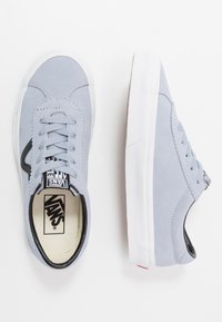 Zapatillas de ante gris con suela de goma blanca, diseño de cordones, detalles en cuero negro y etiqueta de marca en la lengua. Estilo bajo.