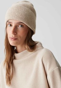 Beanie in maglia a coste beige con un taglio leggermente scivolato, abbinato a un morbido pullover beige chiaro con colletto alto e tessuto testurizzato.