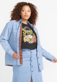 Lichtblauwe denim overhemd gelaagd over een zwarte graphic t-shirt met florale ontwerpen. De outfit heeft een bijpassende denim rok met een split aan de voorkant.