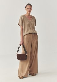 Maglione beige a V con design a treccia, abbinato a pantaloni beige a gamba larga. La modella tiene una borsa in suede marrone scuro con tracolla.