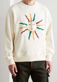 Hellbeiges Sweatshirt mit Sonnenmotiv. Verziert mit einem bestickten Gesicht und mehrfarbigen Strahlen in Grün, Rot, Gelb und Hellblau. Rippbündchen.