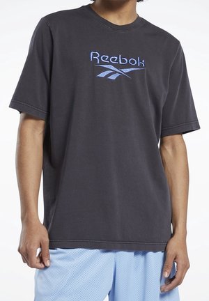 Osoba nosząca ciemnoszarą koszulkę Reebok z niebieskim haftowanym logo i jasnoniebieskie szorty z siateczki, stojąca na białym tle.