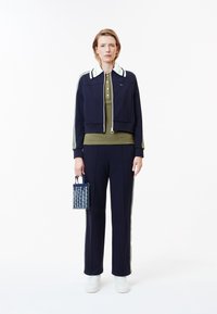 Marinblå kort jacka med gröna och vita detaljer, olivgrön polo, marinblå byxor, vita sneakers och en mönstrad marinblå tote-väska.