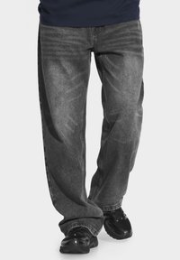 Szerokie szare dżinsy z efektem sprania mają standardowy pięciopunktowy krój, przeszycia oraz są noszone z czarnymi butami sportowymi.