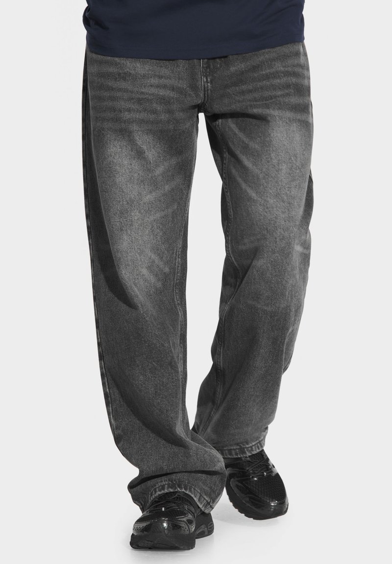 Szerokie szare dżinsy z efektem sprania mają standardowy pięciopunktowy krój, przeszycia oraz są noszone z czarnymi butami sportowymi.