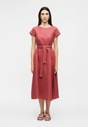 Γυναίκα που στέκεται φορώντας ένα μεσάτο φόρεμα χρώματος σκουριάς με κοντά μανίκια και ζώνη από το ίδιο ύφασμα, σε συνδυασμό με καφέ σανδάλια με επίπεδη σόλα.