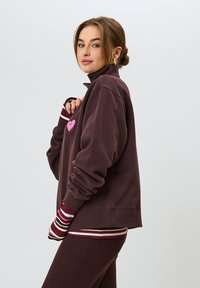 Felpa marrone con colletto alto, grafica a forma di cuore rosa e polsini a righe. Texture morbida e vestibilità ampia, abbinata a pantaloni scuri coordinati.