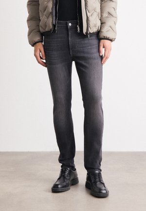 Person trägt schwarze Lederschuhe, dunkle Slim-Fit-Jeans, schwarzen Top und beige gesteppte Jacke und steht auf einem grauen Boden.