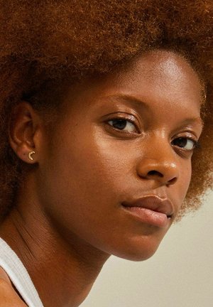 Gros plan sur une personne avec des cheveux bruns naturels et volumineux, portant des boucles d'oreilles en forme de croissant en or, avec des tons de peau chauds et un maquillage subtil.