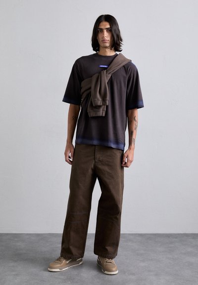 Black oversized t-shirt with blue trim, paired with brown baggy pants and light tan sneakers. A brown sweatshirt draped over one shoulder.