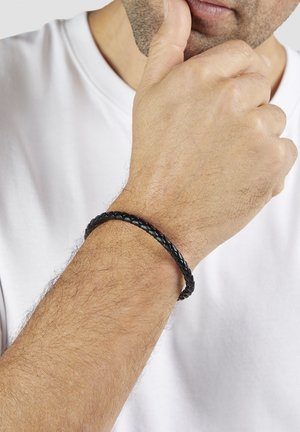 Black braided leather bracelet worn on a wrist, featuring a smooth texture and circular shape, complementing a plain white t-shirt.