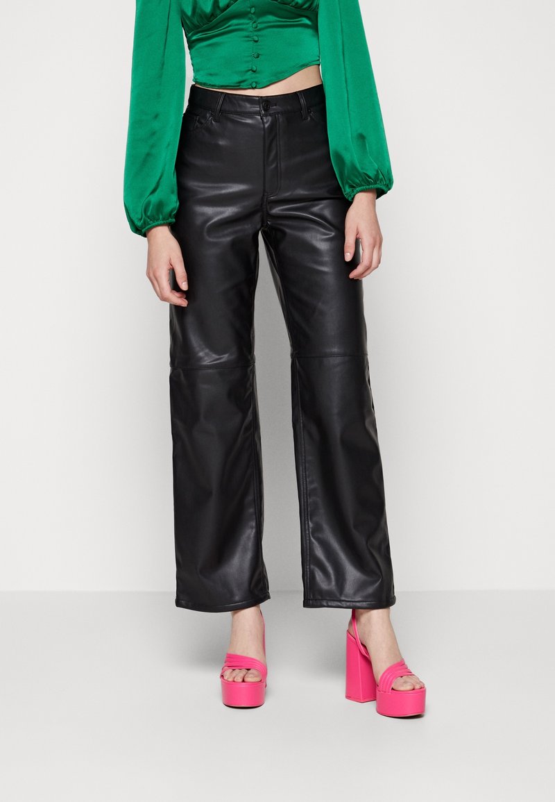 Femme portant un pantalon en cuir noir brillant à jambes larges, un chemisier court à manches longues vert vif et des talons compensés rose fluo.