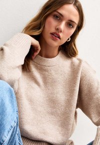 Beżowy sweter dzianinowy z okrągłym dekoltem i ściągaczami, o miękkiej fakturze. Połączenie z niebieskimi dżinsami dopełnia całość.