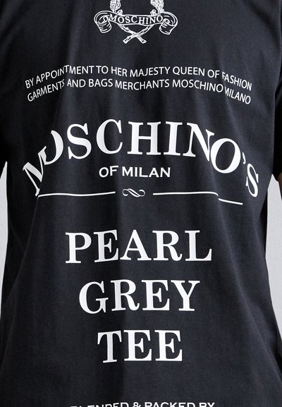 Juodas medvilninis marškinėliai su baltais spausdintais užrašais. Užrašai apima "Moschino's", "Pearl Grey Tee" ir dekoratyvinius elementus. Trumpi rankovės, laisvas kirpimas.