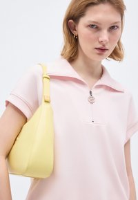 Νεαρή γυναίκα με κοντά ξανθά μαλλιά φοράει ένα ανοιχτό ροζ κοντομάνικο τοπ και κρατά μια ανοιχτό κίτρινη τσάντα ώμου στο αριστερό της χέρι.