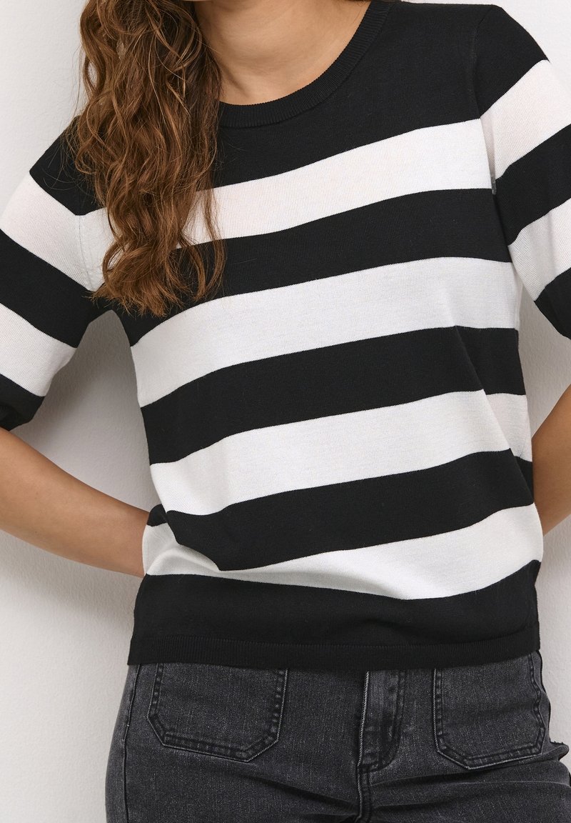 Femme portant un pull rayé noir et blanc à manches courtes, avec un jean gris foncé, les mains sur les hanches, les cheveux sur une épaule.