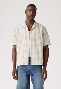 Korte mouwen overhemd met een beige patroonstof met kleine blauwe en groene ruiten, gecombineerd met lichte denim jeans.