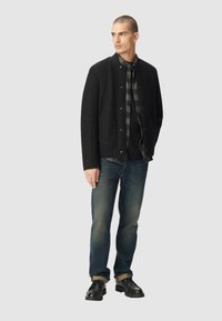 Czarna kurtka z mieszanki wełny z zatrzaskami, zestawiona z ciemną kraciastą koszulą, niebieskimi dżinsami i czarnymi skórzanymi butami. Model stoi na neutralnym tle.