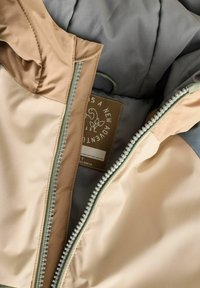 Veste légère et rembourrée avec des panneaux beige et gris, fermetures éclair contrastantes et une étiquette d'entretien avec un logo de plante à l'intérieur.