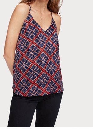 Vrouw met een mouwloze V-hals top met een blauw-rood geometrisch vierkant patroon en zwarte broek, staand tegen een effen achtergrond.