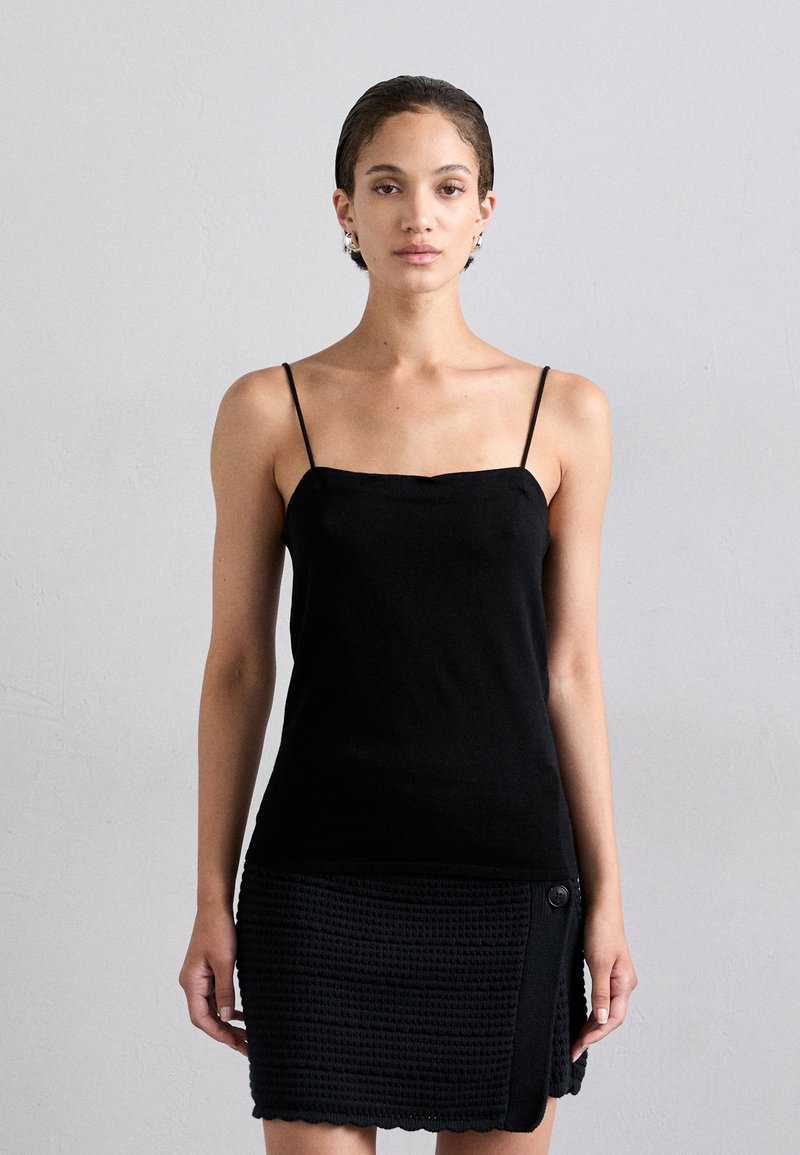 Black sleeveless top with thin straps, featuring a fitted design and smooth fabric, paired with a textured black skirt with a button detail.