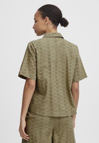 Olive green short-sleeved shirt featuring a floral eyelet pattern, soft texture, and a classic collar, worn with matching shorts.