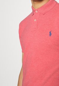 Kurzarm-Polohemd in leuchtendem Koralle, aus strukturiertem Baumwollmaterial, mit einer Drei-Knopf-Leiste und einem blauen gestickten Logo auf der Brust.