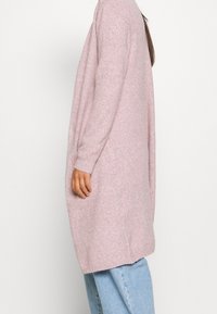 Donna che indossa un lungo cardigan a maglia morbida di colore rosa chiaro sopra jeans azzurri, in piedi di lato contro uno sfondo bianco semplice.