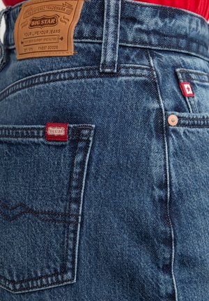 Tmavě modré džíny z denimu s texturovaným povrchem. Mají světle hnědou koženou nášivku, červenou logo visačku a viditelné švy na kapsách.