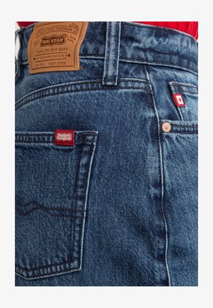 Tamsiai mėlyni džinsai su tekstūruotu paviršiumi. Yra rudas odinis ženklas, raudonas logotipo etiketas ir matomi siūlų akcentai ant kišenių.