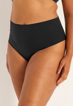 Close-up van een persoon die hooggeknipte zwarte onderbroek draagt, hand rustend op het bovenbeen, zichtbaar bovenlichaam en bovenbenen.