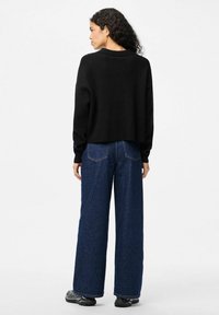 Črni kratki pulover z dolgimi rokavi, kombiniran z širokimi modrimi kavbojkami, ki imajo teksturirano tkanino in vidne zadnje žepe. Videz dopolnjujejo superge.