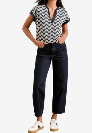 Vrouw met een kortere mouw navyblauw en wit patroon V-hals top, donkerblauwe high-waist jeans en witte sneakers met bruine zolen, staand met handen in de zakken.