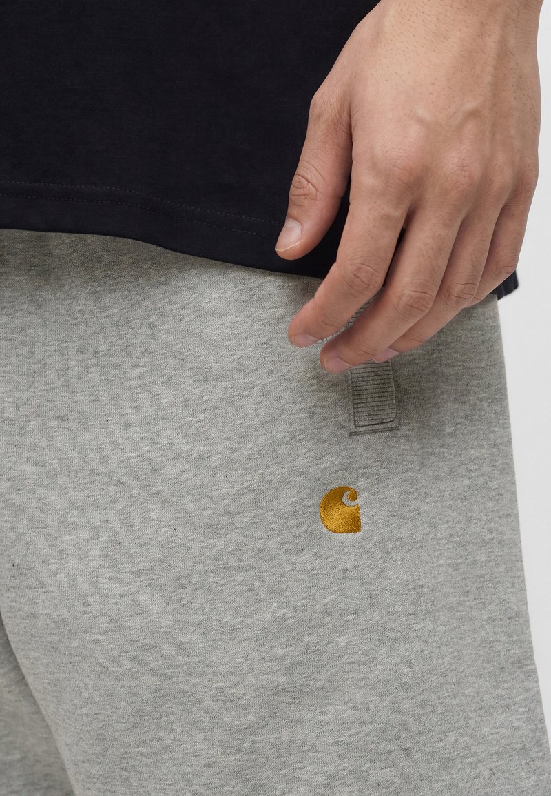 Hand ruht in der Nähe der Tasche auf hellgrauen Jogginghosen mit kleinem gelben Logo, kombiniert mit schwarzem Shirt.