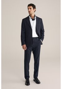 Uomo con abito blu navy, camicia bianca, sciarpa al collo nera con motivo, scarpe eleganti nere, in piedi con una mano in tasca.