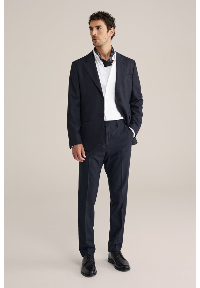 Uomo con abito blu navy, camicia bianca, sciarpa al collo nera con motivo, scarpe eleganti nere, in piedi con una mano in tasca.