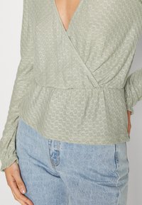 Top envolvente texturizado em verde claro, com cintura peplum, mangas longas e decote em V profundo, combinado com jeans azuis de cintura alta.