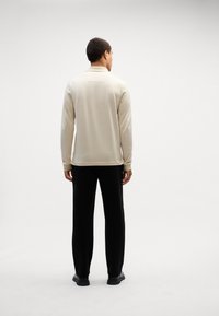 Lys beige langermet polo skjorte med teksturert mønster, ryggdetalj, kombinert med svarte bukser og svarte sko.