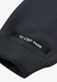 Dunkelgrauer Sweatshirtstoff mit gerippten Bündchen. Das Etikett zeigt "ICI C'EST PARIS" in Weiß auf schwarzem Hintergrund. Glatte Textur, lässiges Design.