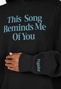 Čierna mikina vyrobená z mäkkej látky, s svetlomodrým textom "Táto pieseň mi pripomína teba" na prednej strane a "Pegador" na rukáve.