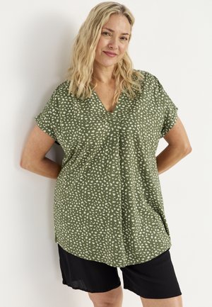 Lyshåret kvinde stående med hænderne bag ryggen, iført grøn mønstret bluse og sorte shorts, foran en hvid baggrund.