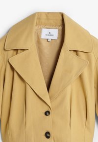 Cappotto in tessuto giallo intrecciato con colletto a revers, chiusura con bottoni e dettagli plissettati; l'interno presenta un rivestimento morbido con motivi sottili.